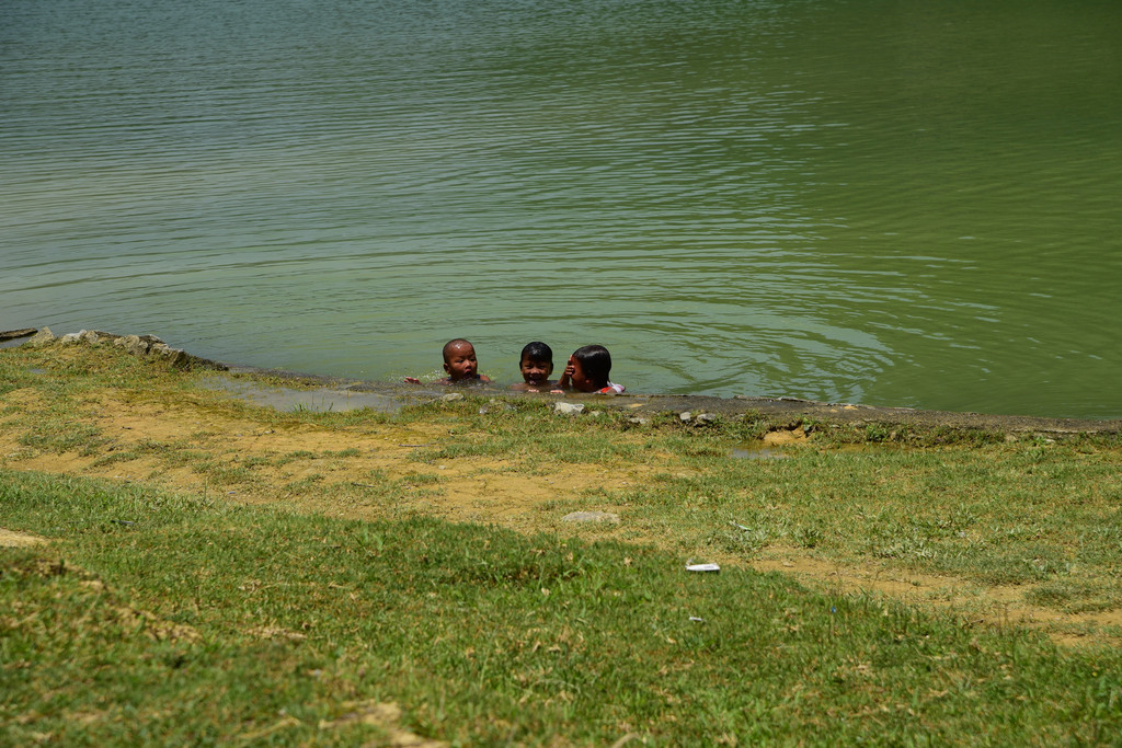 蓝色衣服的女人在湖边一棵大树下纳凉闲聊,小孩光着屁股在湖里戏水,一